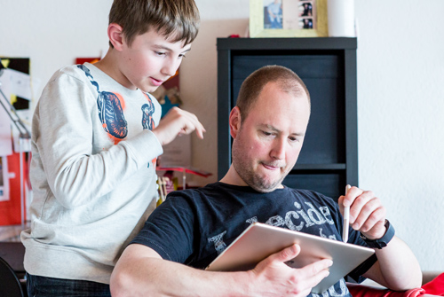 Vater Lukas erklärt Janis etwas an einem Tablet.